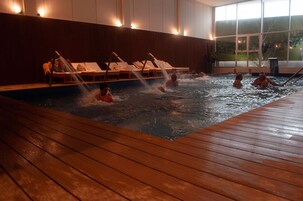 Piscina cubierta climatizada con plataforma de madera, sillas de descanso y vistas al exterior a través de amplios ventanales.