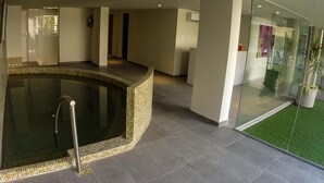 Un área de piscina interior moderna con borde de azulejos, un muro de cristal con césped artificial verde y un extintor visible en la pared.