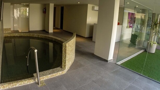 A modern indoor pool area with a tiled edge, a glass wall with a green artificial lawn, and a visible fire extinguisher on the wall.