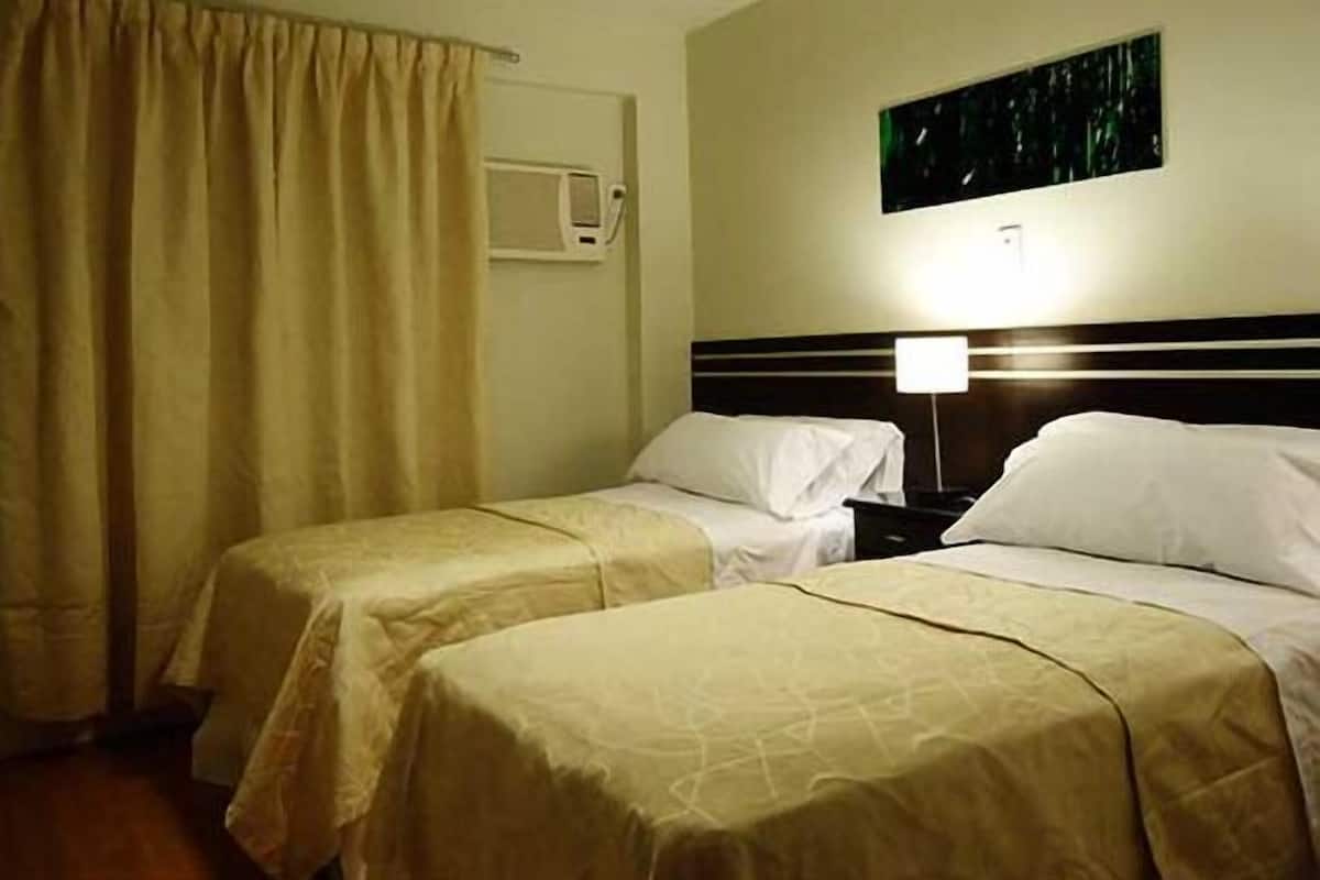 A hotel room with two beds, a bedside table with a lamp, a window with curtains, and a wall-mounted air conditioning unit.