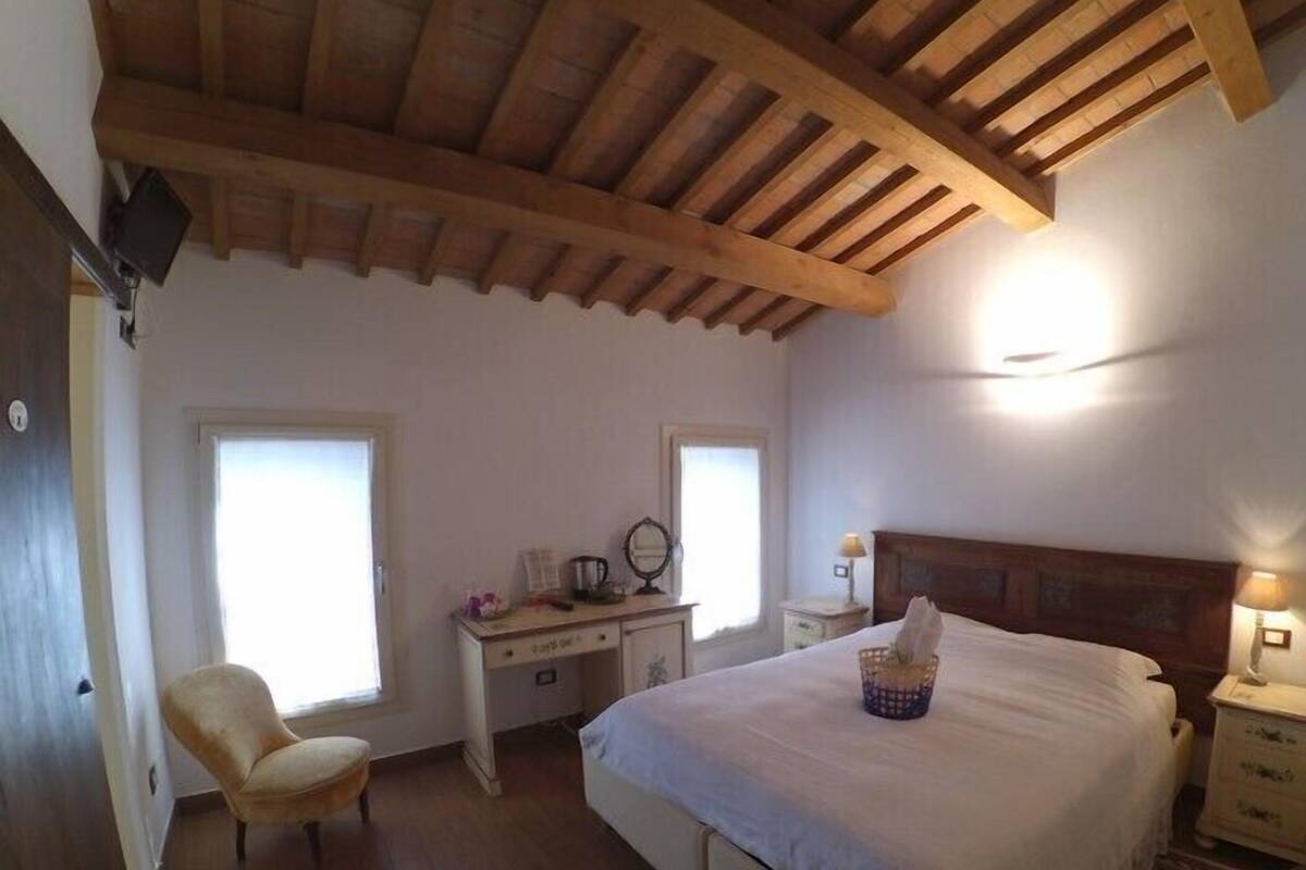 A bedroom with a wooden ceiling, a bed with a white bedspread, a desk with a chair, and a nightstand with a lamp.