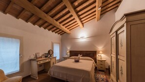 Un dormitorio con techo de madera, una cama con colcha blanca, una mesita de noche con lámpara, un aparador y una ventana con persianas.