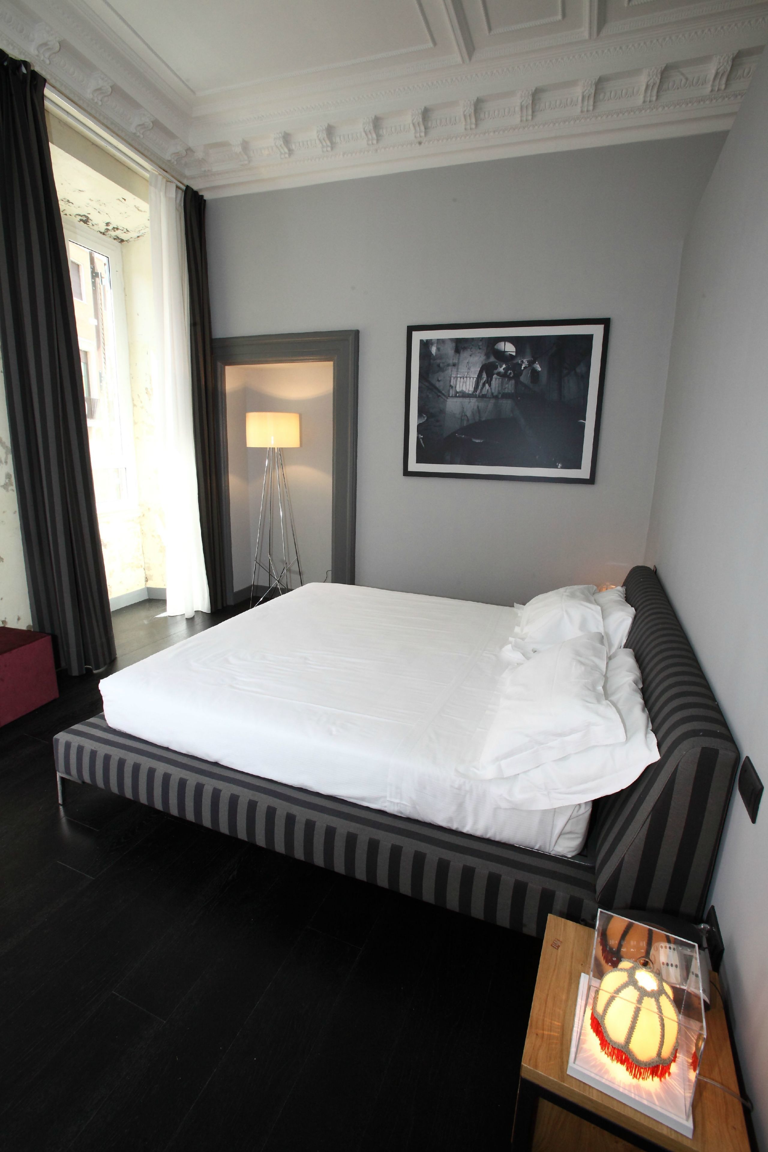 A hotel room with a large bed, a bedside table with a lamp, a framed picture on the wall, and a view of the outdoors through a window.
