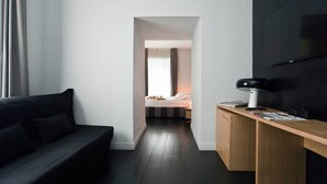 Quarto de hotel moderno com piso de madeira escura, sofá preto, escrivaninha de madeira com computador e cama com cabeceira branca.