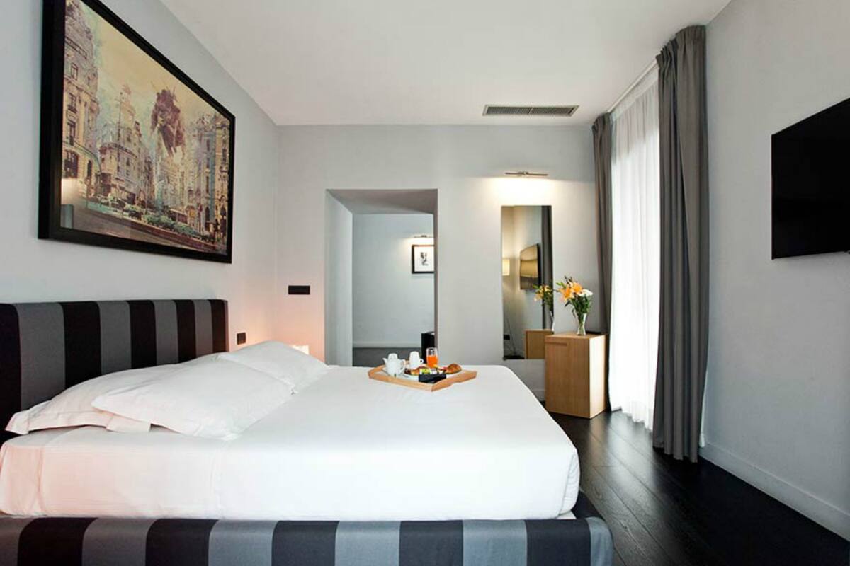 A hotel room with a large bed, a painting on the wall, a bedside table with a tray of breakfast items, a mirror, and a TV.