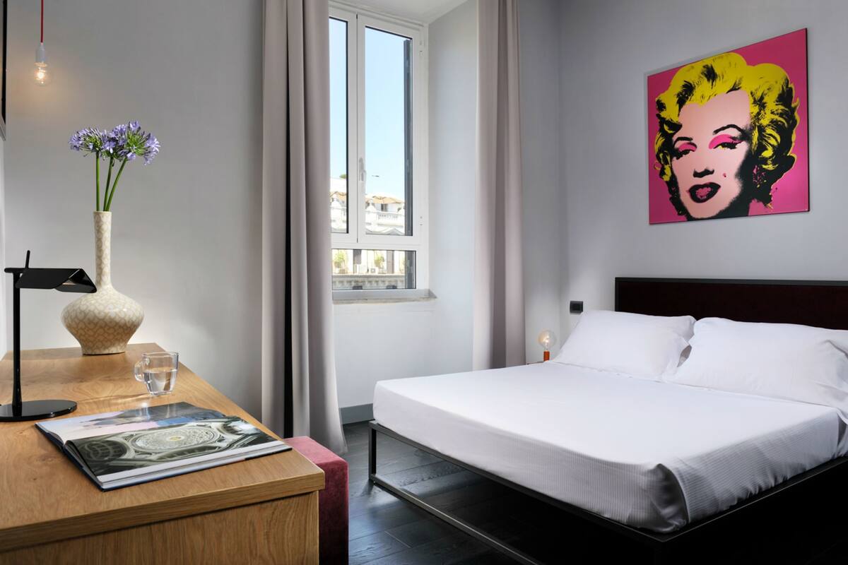 A hotel room with a bed, a bedside table with a lamp, a vase with flowers, and a window with curtains.