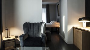 Quarto de hotel moderno com uma poltrona cinza, uma mesa de cabeceira com abajur, uma cama com roupa de cama branca e um armário de madeira.