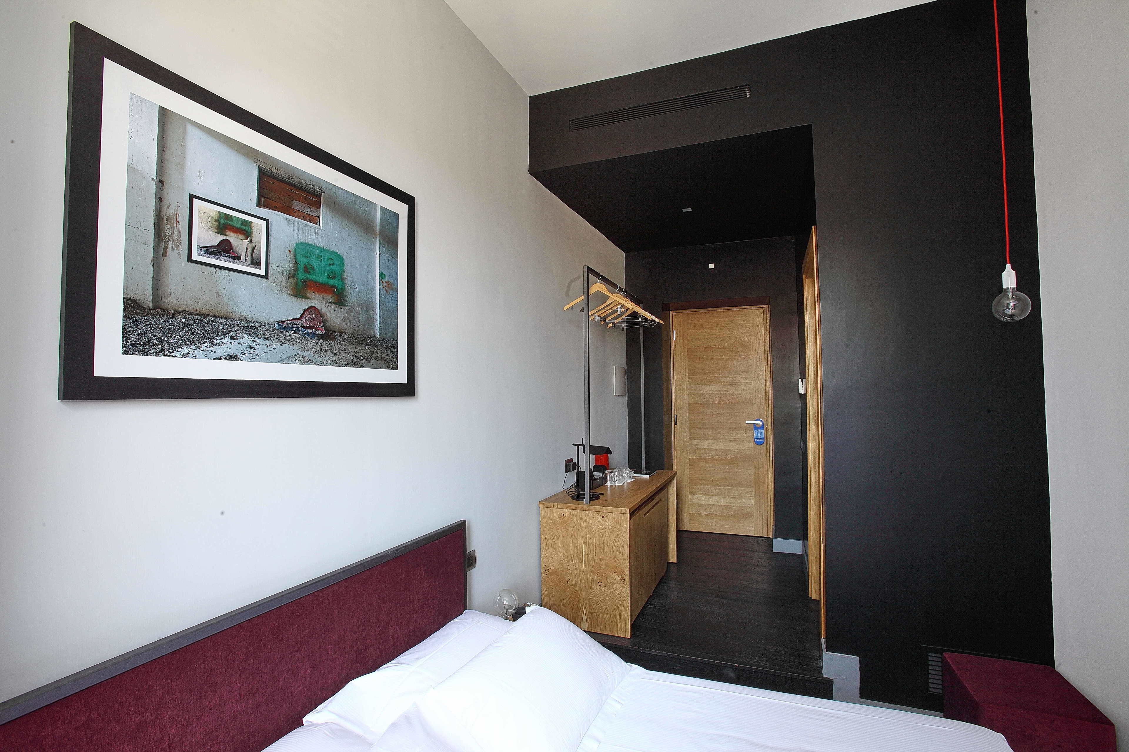 A hotel room with a bed, a wooden desk, a mirror, and a framed picture on the wall.
