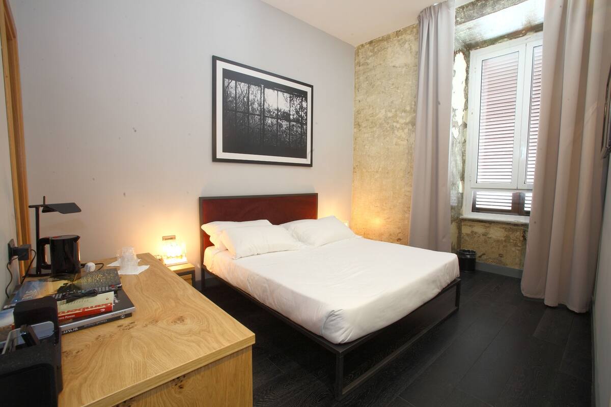 A bedroom with a bed, a desk with books, a framed picture on the wall, and a window with curtains.