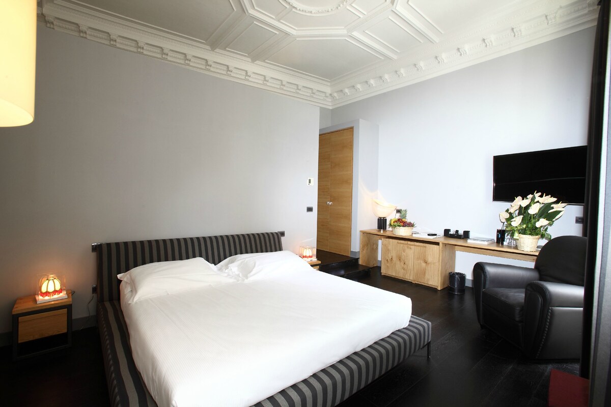 A hotel room with a large bed, a desk, a chair, a TV, and a nightstand with a lamp.
