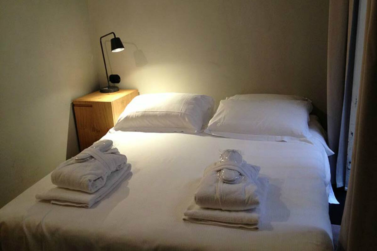 A neatly made bed with white linens and two folded towels on the bed. There is a small wooden nightstand with a black lamp beside the bed.