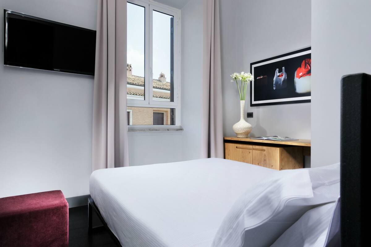 A hotel room with a bed, a TV mounted on the wall, a window with a view of buildings, a wooden desk, and a vase with flowers.