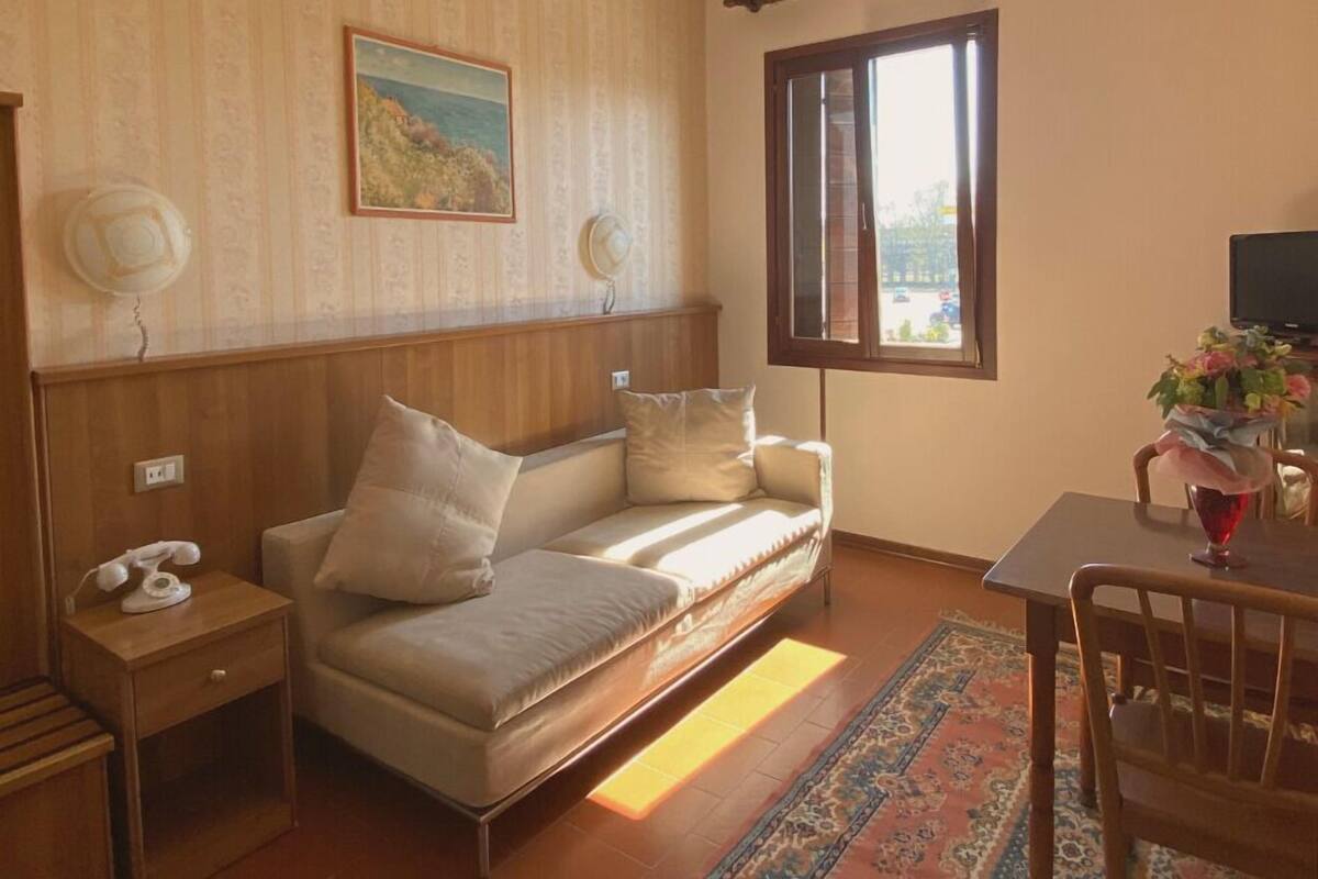 Un cuarto acogedor con un sofá, una mesita con un florero, una silla, un cuadro en la pared y una ventana con vistas.