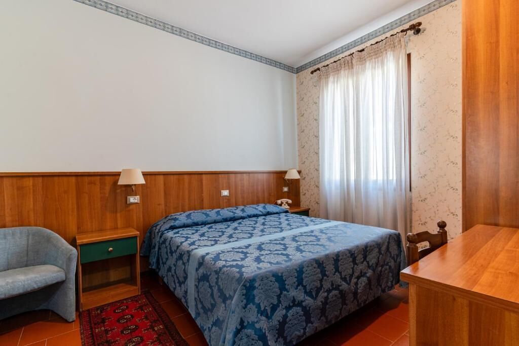 Un dormitorio con cama, una silla, una mesita de noche, una lámpara y una mesa de madera.