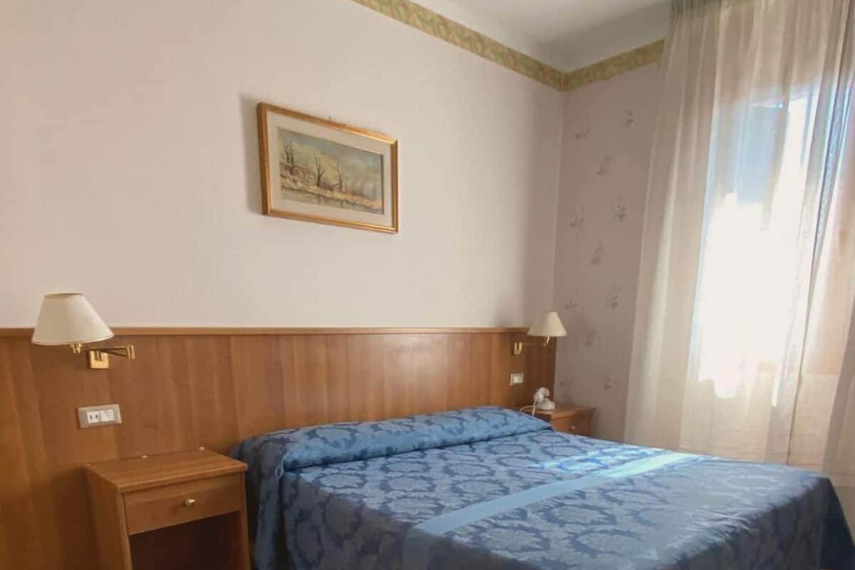 Habitación de hotel con cama, dos mesitas de noche, una lámpara, un cuadro en la pared y una ventana con cortinas.