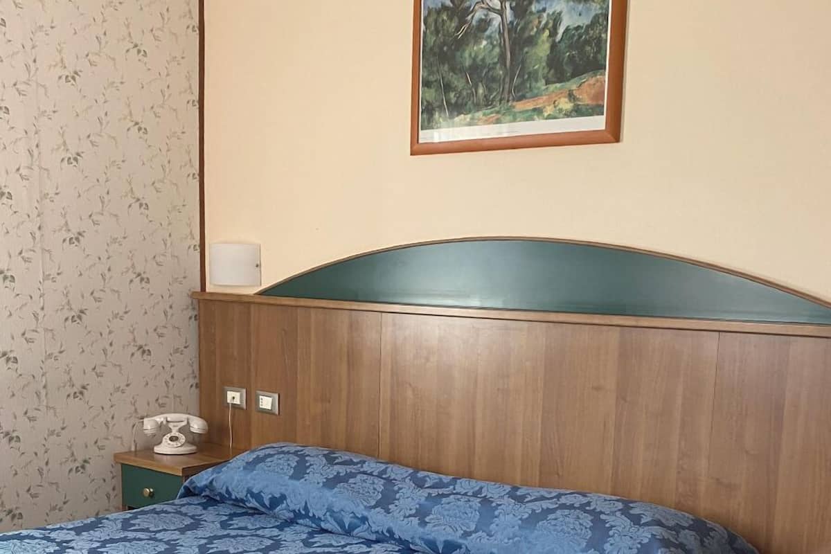 Una habitación con cabecera de madera, cama con colcha azul estampada, papel tapiz floral, un cuadro de un árbol y un teléfono antiguo sobre una mesita de noche.