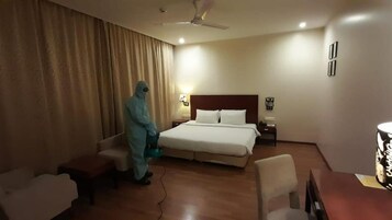 A hotel room with a bed, a desk, a chair, a nightstand, a lamp, and a person in a protective suit.