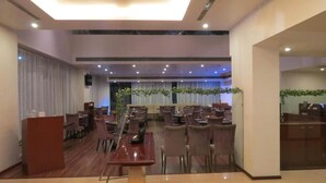 A hotel dining area with multiple tables and chairs, a podium, and a large window with curtains. - The Aures (Chhatrapati Sambhajinagar)