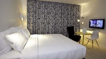 Moderni hotellihuone, jossa on suuri sänky, kuviollinen pääty, pieni pyöreä pöytä ja tuoli, televisio sekä seinään kiinnitetty valaisin.