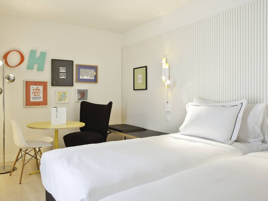 Ein Hotelzimmer mit einem Bett, einem kleinen Tisch, einem Stuhl und einer Wand mit gerahmten Bildern und einem Schild.