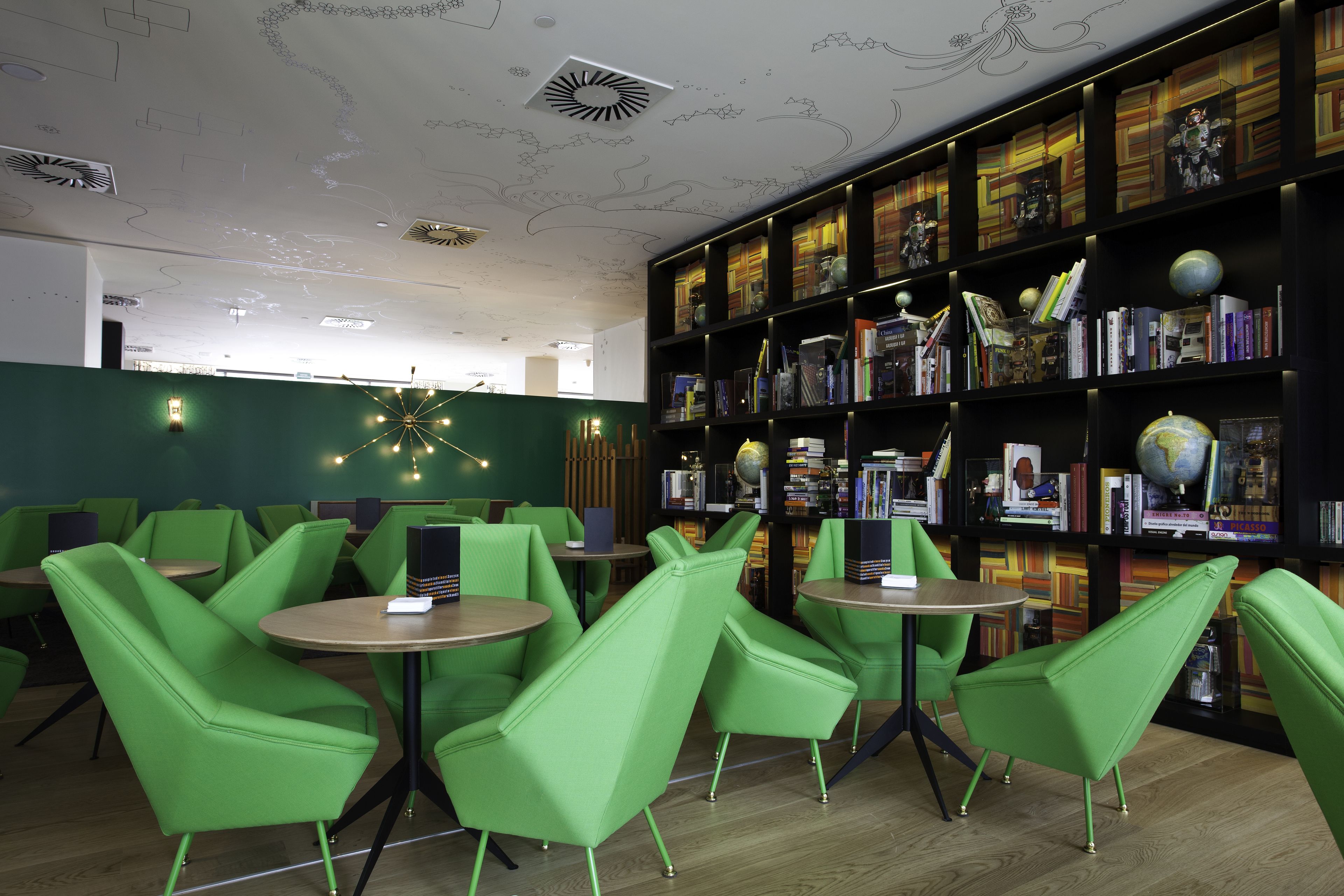 A modern library with green chairs and tables, a large bookshelf with books and decorative items, and a unique ceiling design.