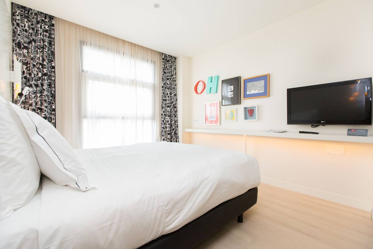 Moderni hotellihuone, jossa on suuri sänky, taulutelevisio ja ikkuna, jossa on läpikuultavat verhot.