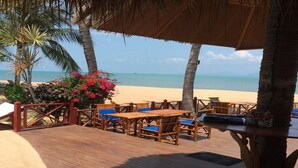 Una terraza de madera con mobiliario exterior, techo de paja y vista a la playa y al océano.