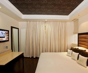 A hotel room with a large bed, a TV on the wall, and a curtain. - Madhuban Hotel (New Delhi)