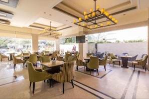 A spacious indoor area with multiple tables and chairs, large windows, and a modern chandelier. - Madhuban Hotel (New Delhi)
