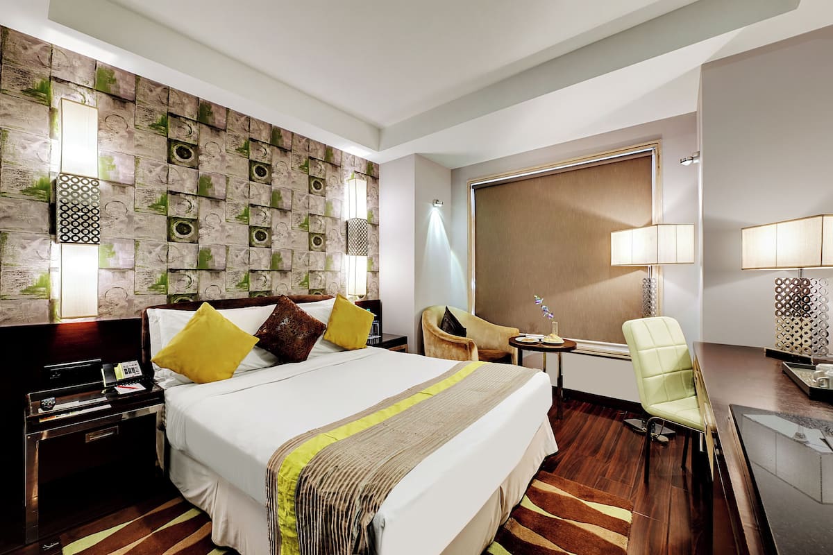 A hotel room with a large bed, a desk with a chair, a wall-mounted TV, and a window with blinds.