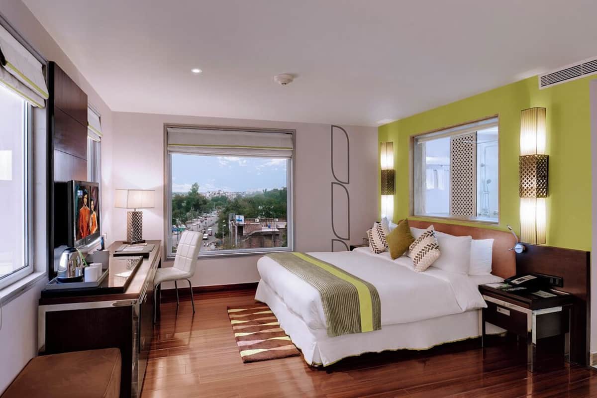 A modern hotel room with a large bed, a desk with a computer, a chair, and a view of the city.