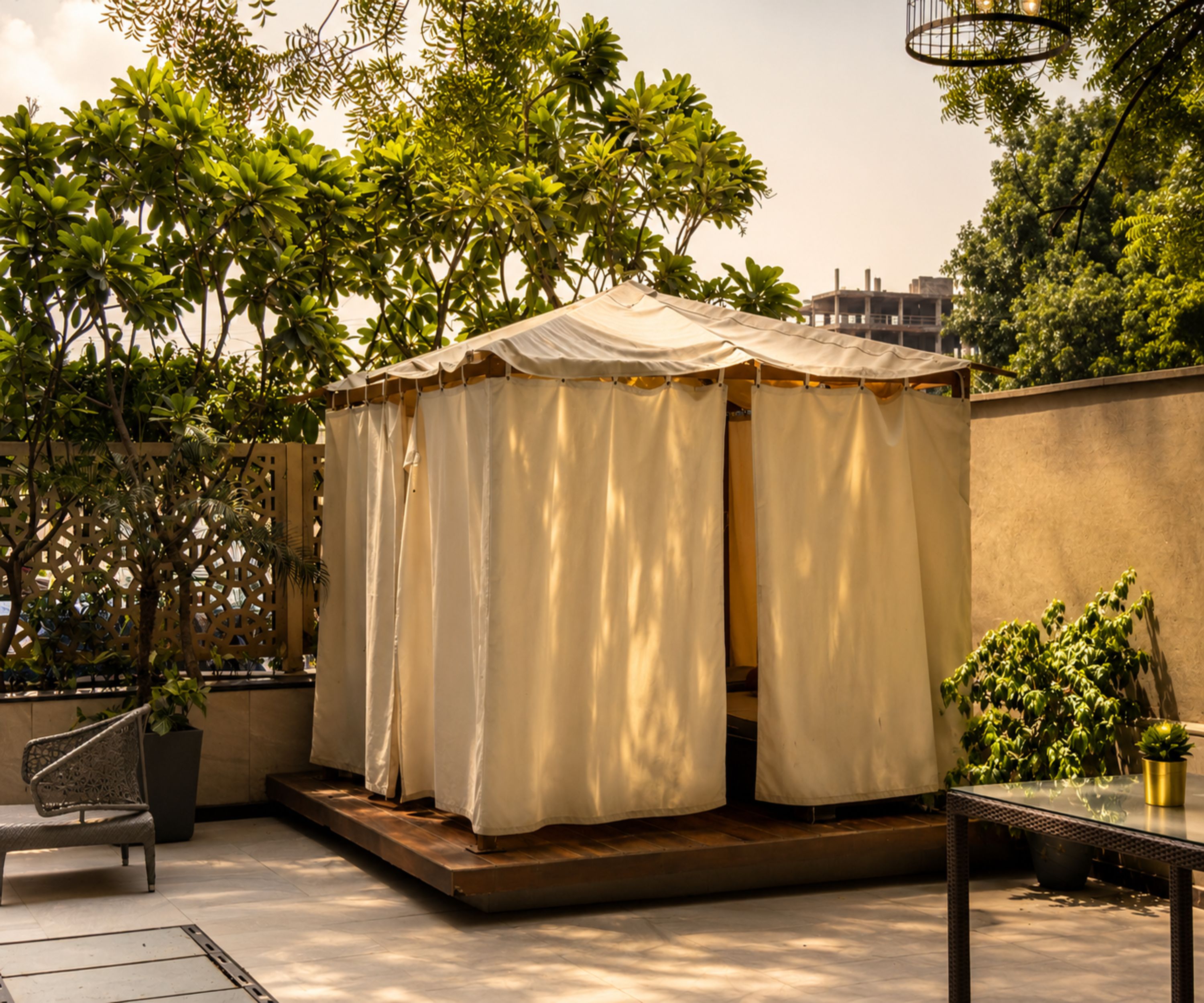 A covered outdoor seating area with a table and chairs, surrounded by greenery and a building in the background.