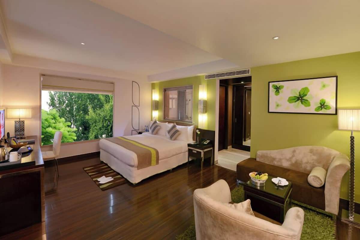A hotel room with a bed, a desk with a lamp, a chair, a sofa, and a painting on the wall.