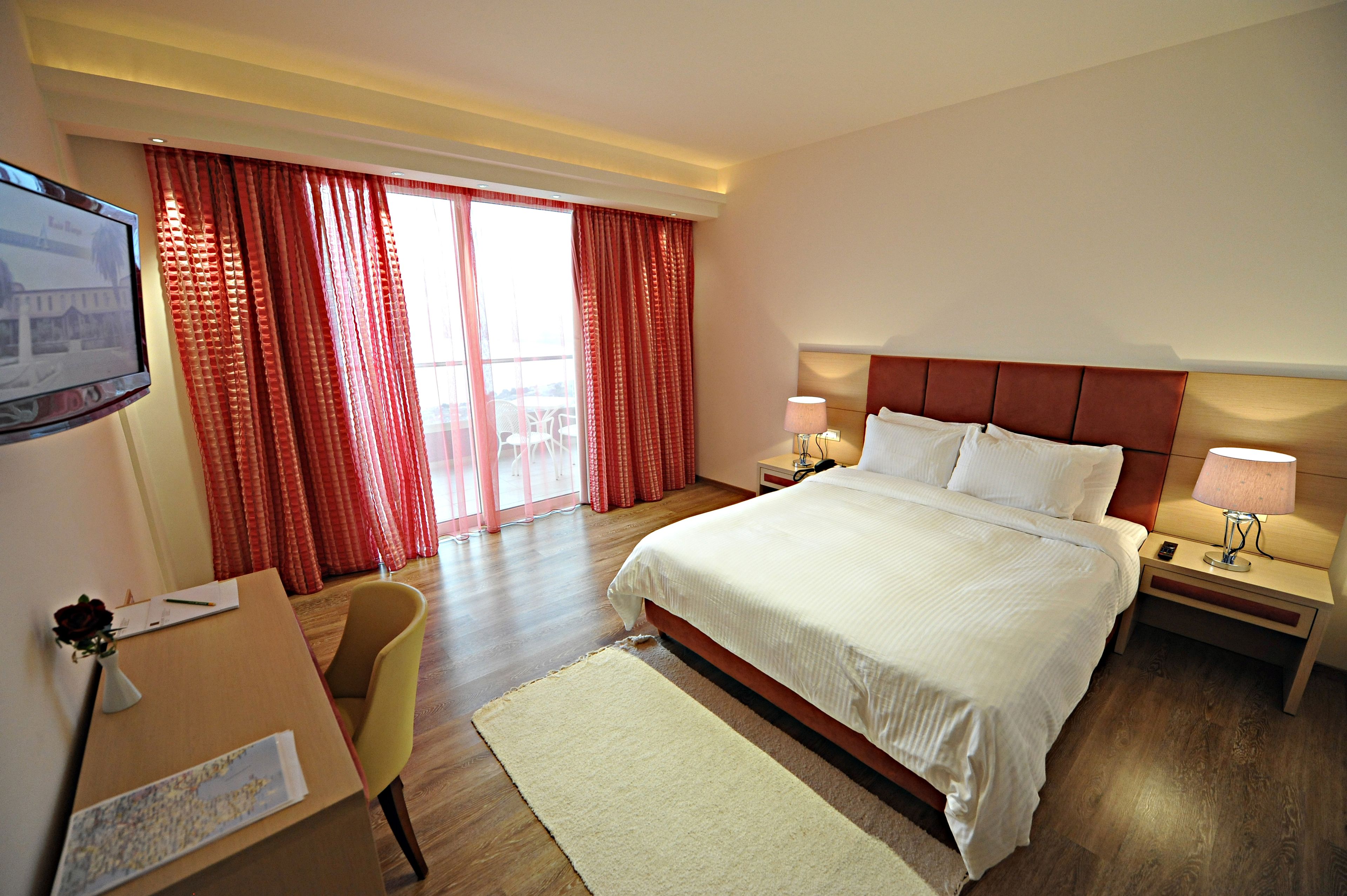 A hotel room with a large bed, a desk with a chair, a TV, and a window with red curtains.