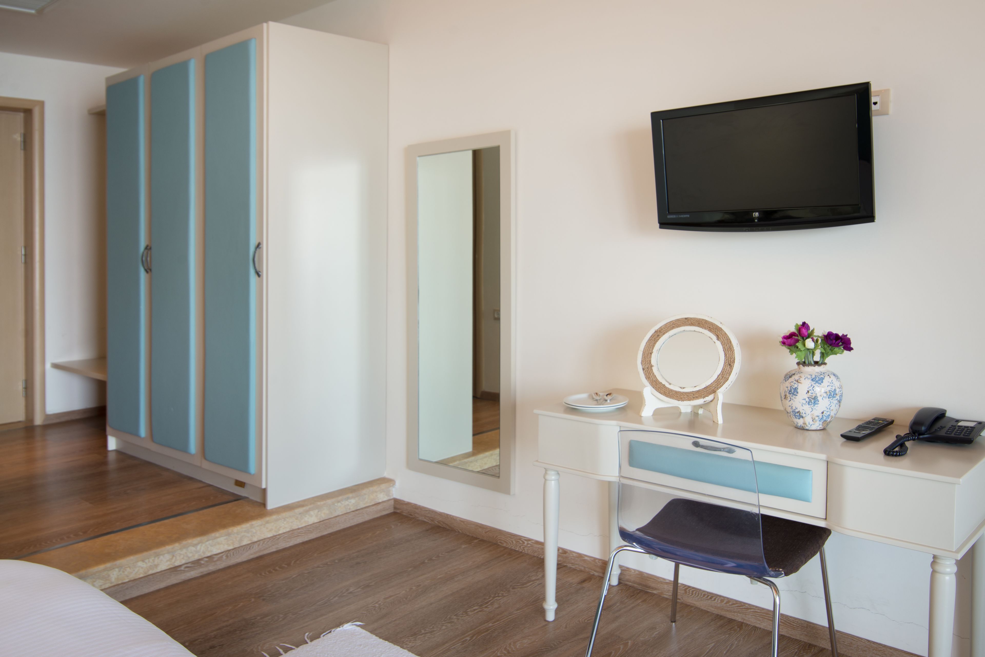 A bedroom with a bed, a desk with a mirror, a vase with flowers, a telephone, and a television on the wall.