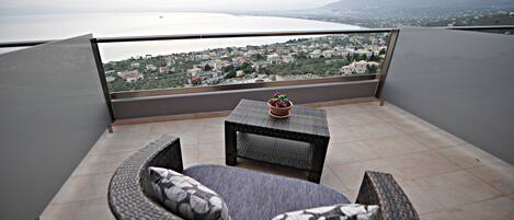 A balcony with wicker furniture, a small table with a potted plant, and a view of a coastal town and mountains.