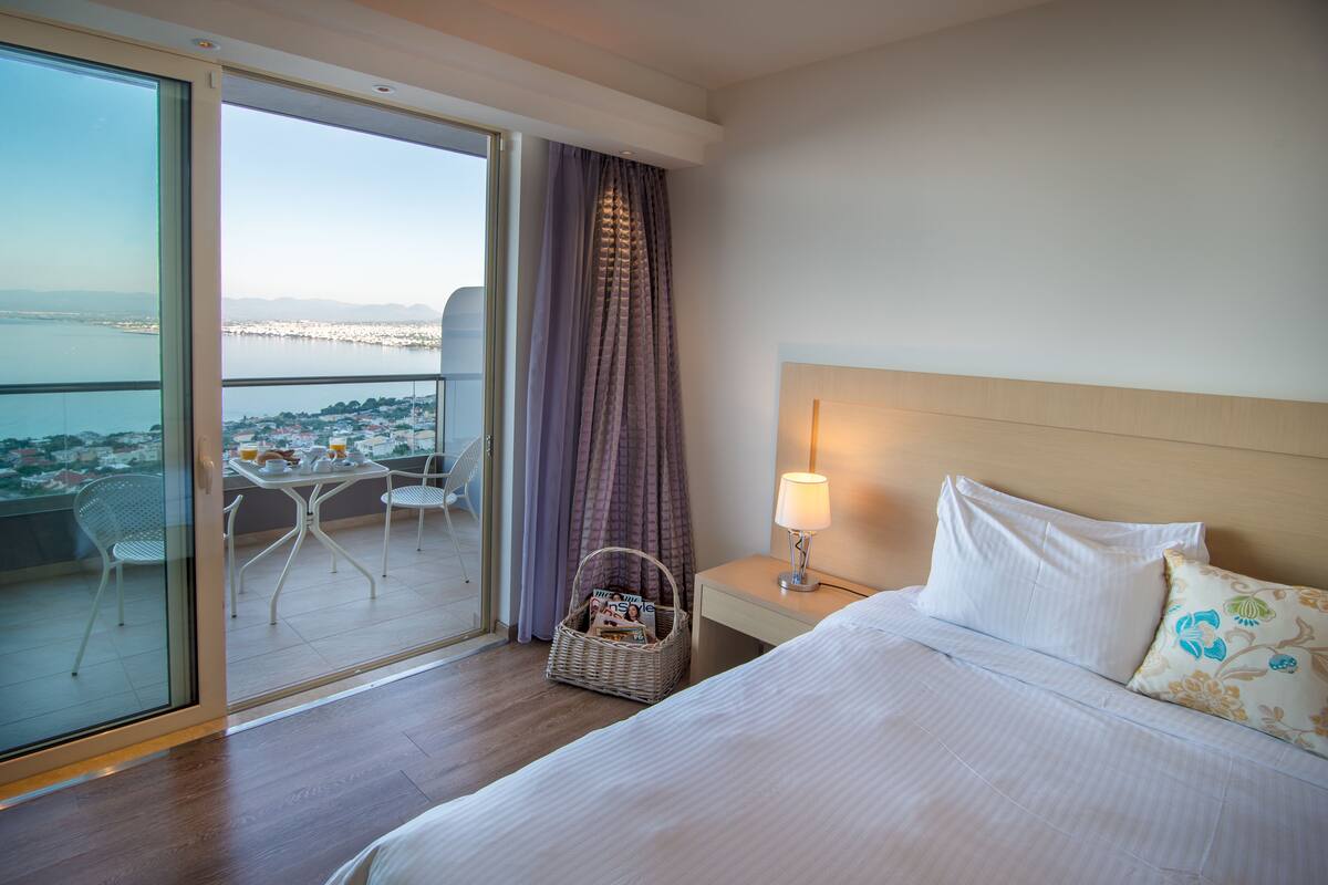 Une chambre d’hôtel comprenant un lit, une table de chevet, une lampe, un panier en osier, une table avec des chaises, et une vue sur la ville et l’eau, visible à travers une porte vitrée.