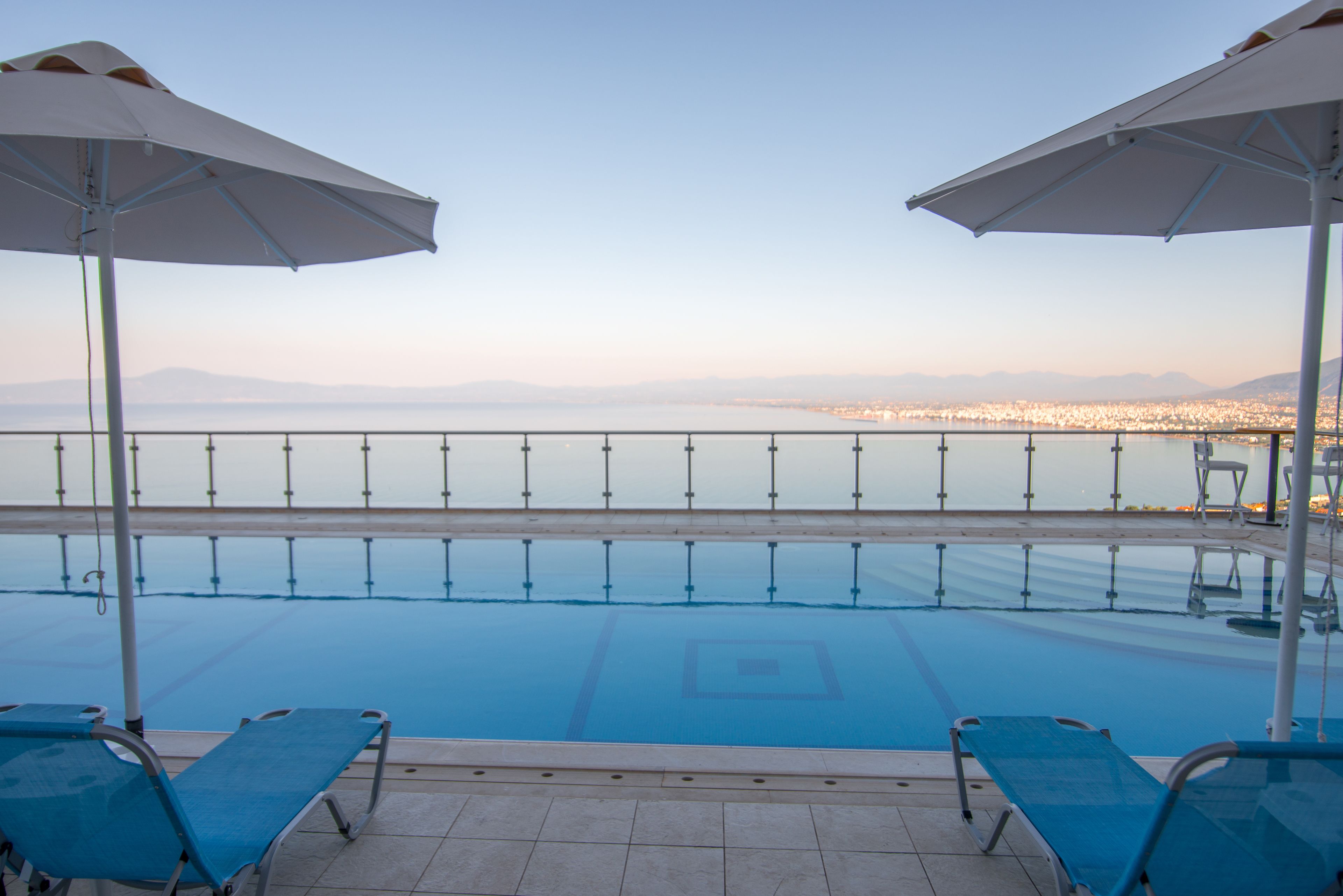 A poolside area with two blue lounge chairs and two white umbrellas, overlooking a calm body of water and a distant cityscape.