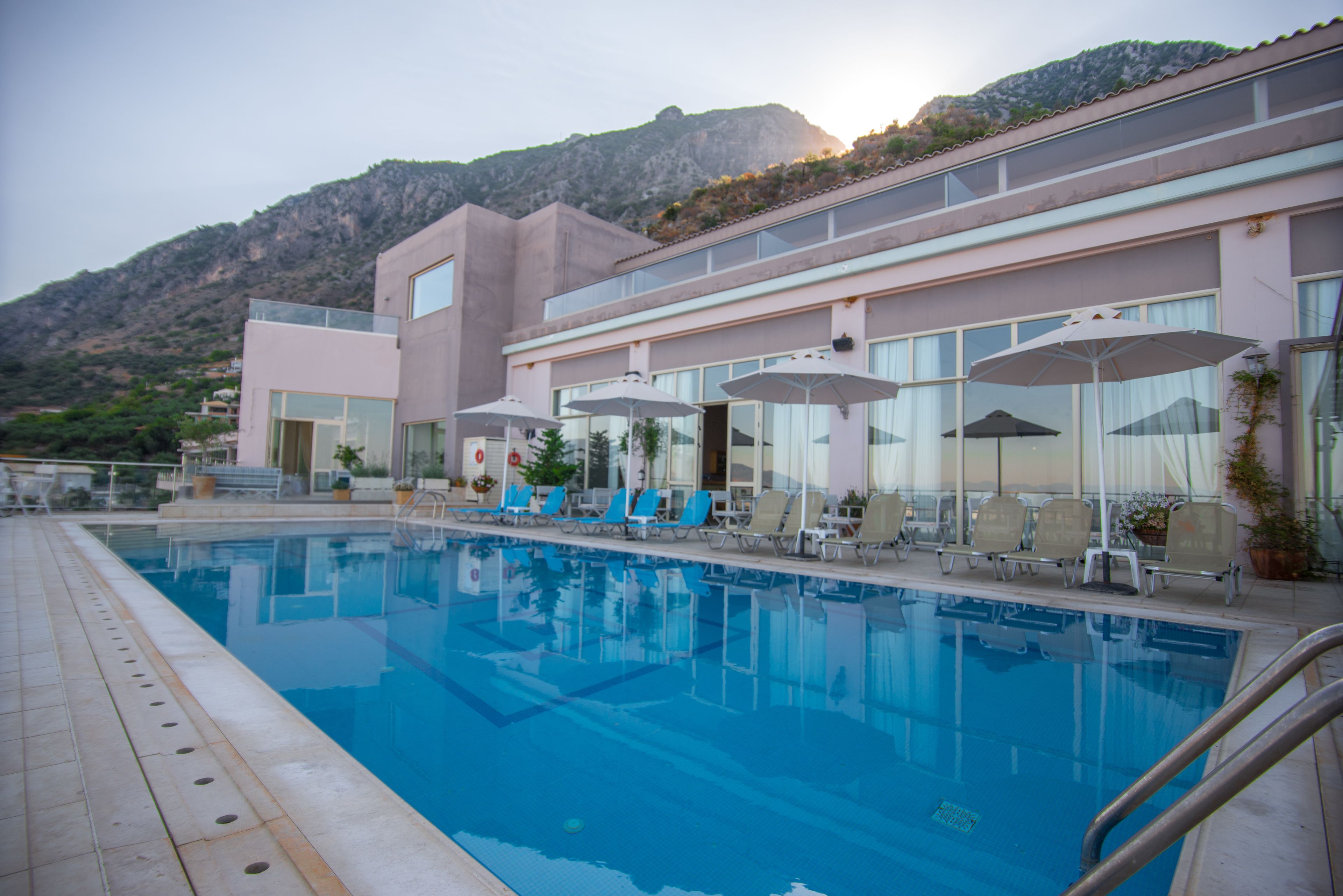 A swimming pool with lounge chairs and umbrellas, a building with large windows, and a mountainous backdrop.
