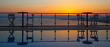An infinity pool with a sunset view, outdoor seating with chairs and tables, and a railing.