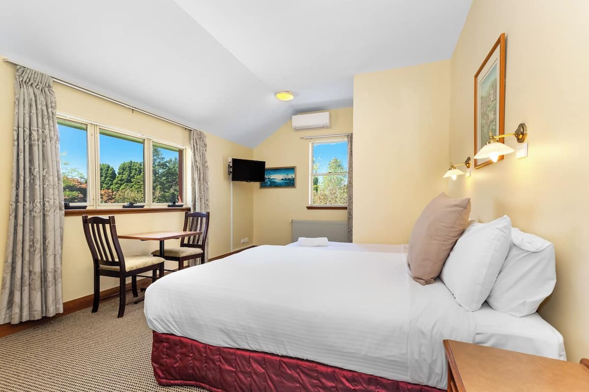 A hotel room with a large bed, a desk, and two chairs. There are windows with curtains, a flat-screen TV, and wall-mounted lights.