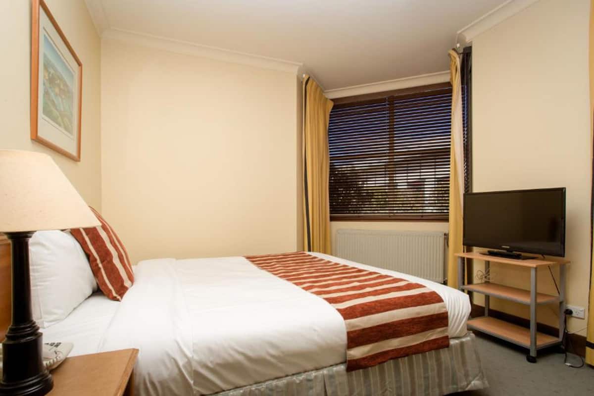 A hotel room with a bed, a TV on a stand, a lamp, and a window with blinds.