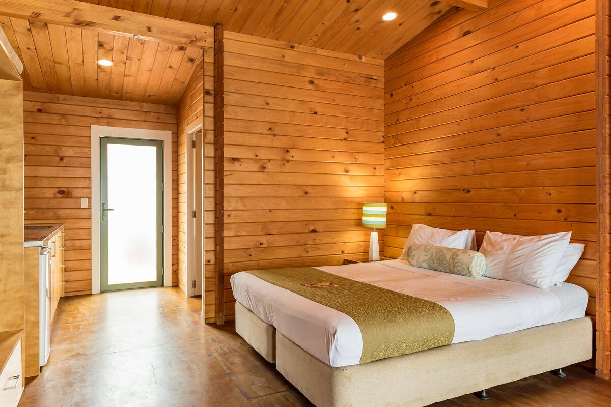 A wooden-paneled room with a bed, a bedside lamp, and a door leading to another room.