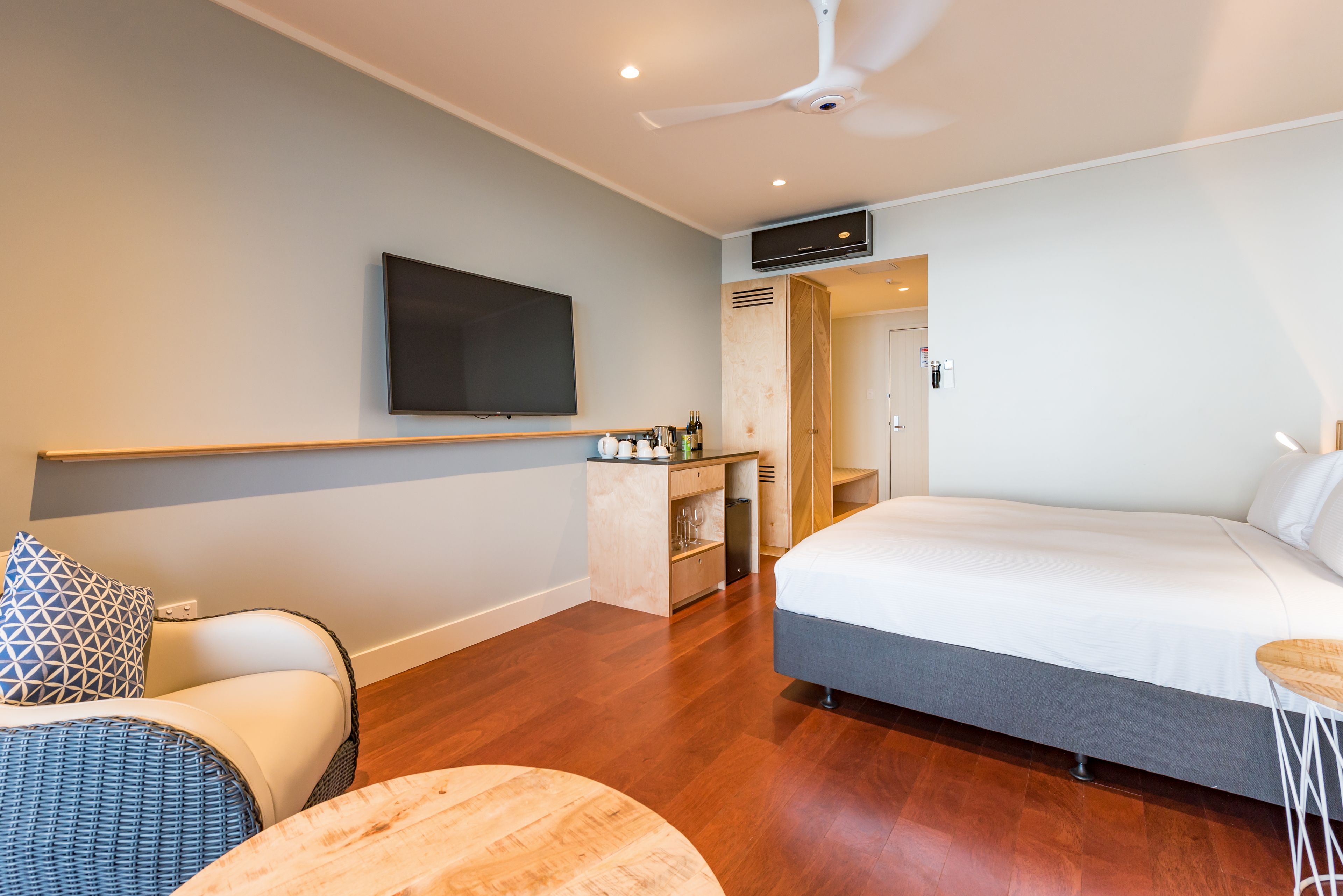 Una habitación de hotel moderna con una cama grande, un televisor de pantalla plana montado en la pared, un pequeño rincón de descanso con una silla y reposapiés, y un mueble de madera con botellas y vasos.