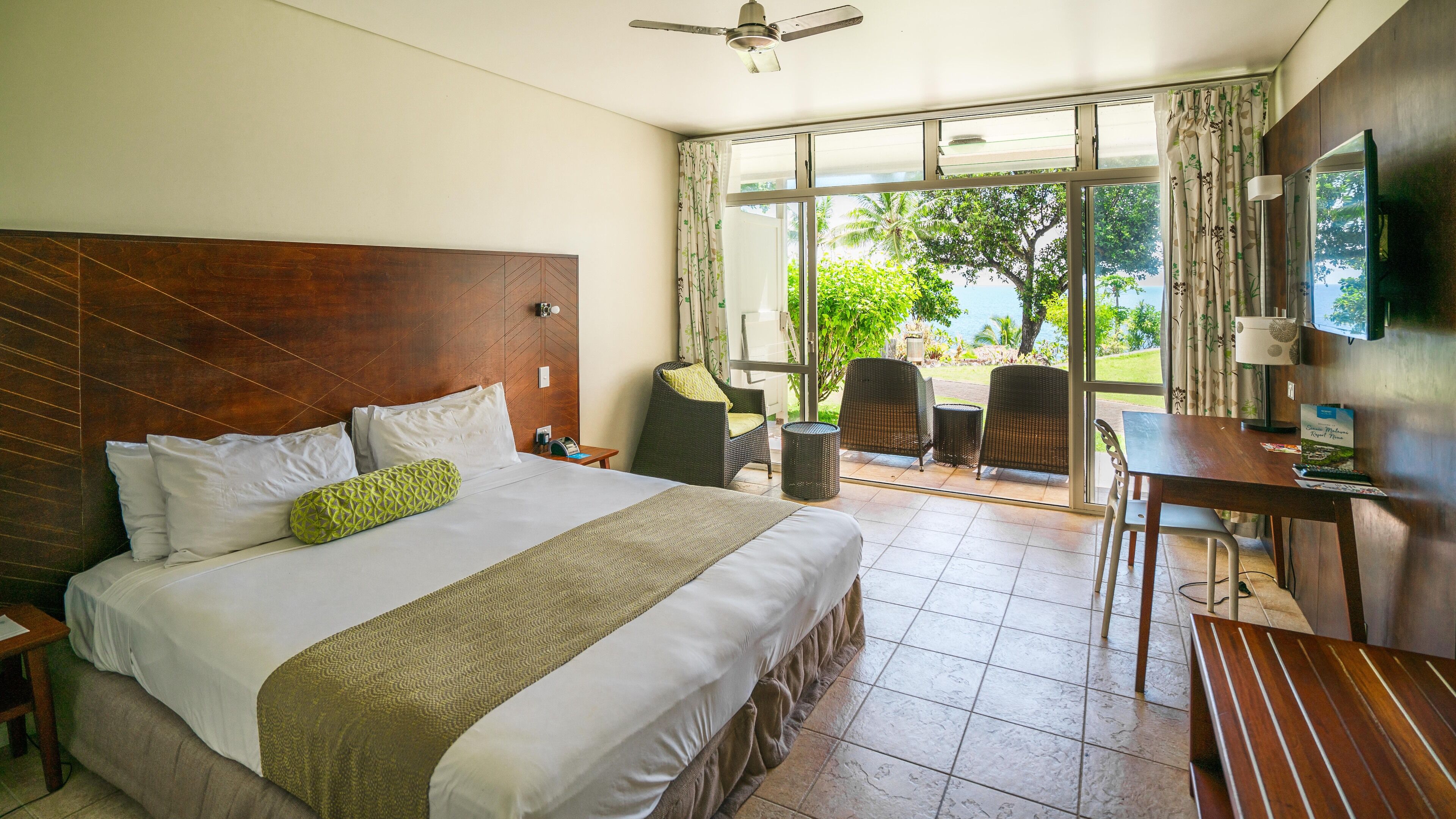 Ein Hotelzimmer mit einem großen Bett, einem Schreibtisch und einem Stuhl. Ein Fenster bietet Blick auf Grünflächen und eine Palme.