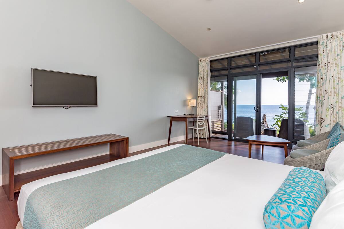 A hotel room with a large bed, a TV mounted on the wall, a desk with a chair, and a view of the ocean through large sliding doors.