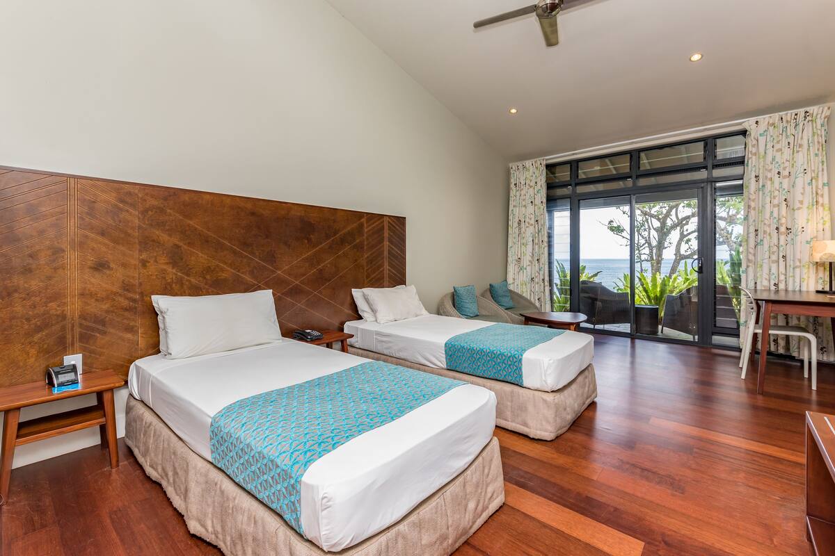 A hotel room with two beds, a wooden headboard, a desk, and a view of the ocean through large windows.