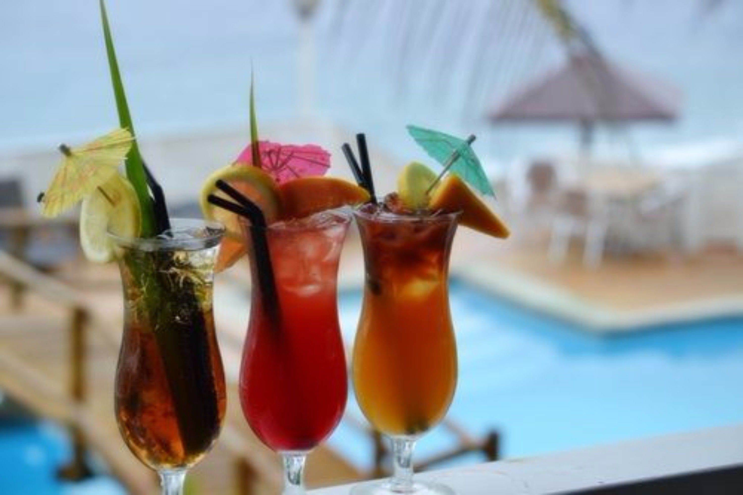 Three cocktails with garnishes on a table near a pool.