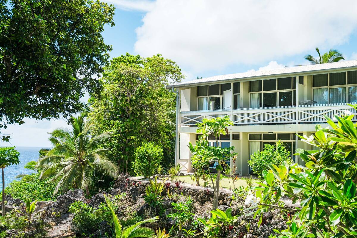 A two-story building with a balcony, surrounded by tropical vegetation and overlooking a body of water.