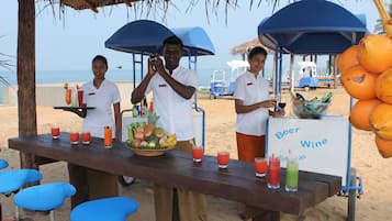 Un bar frente a la playa con una mesa de madera que muestra diversas bebidas y una cesta de frutas.